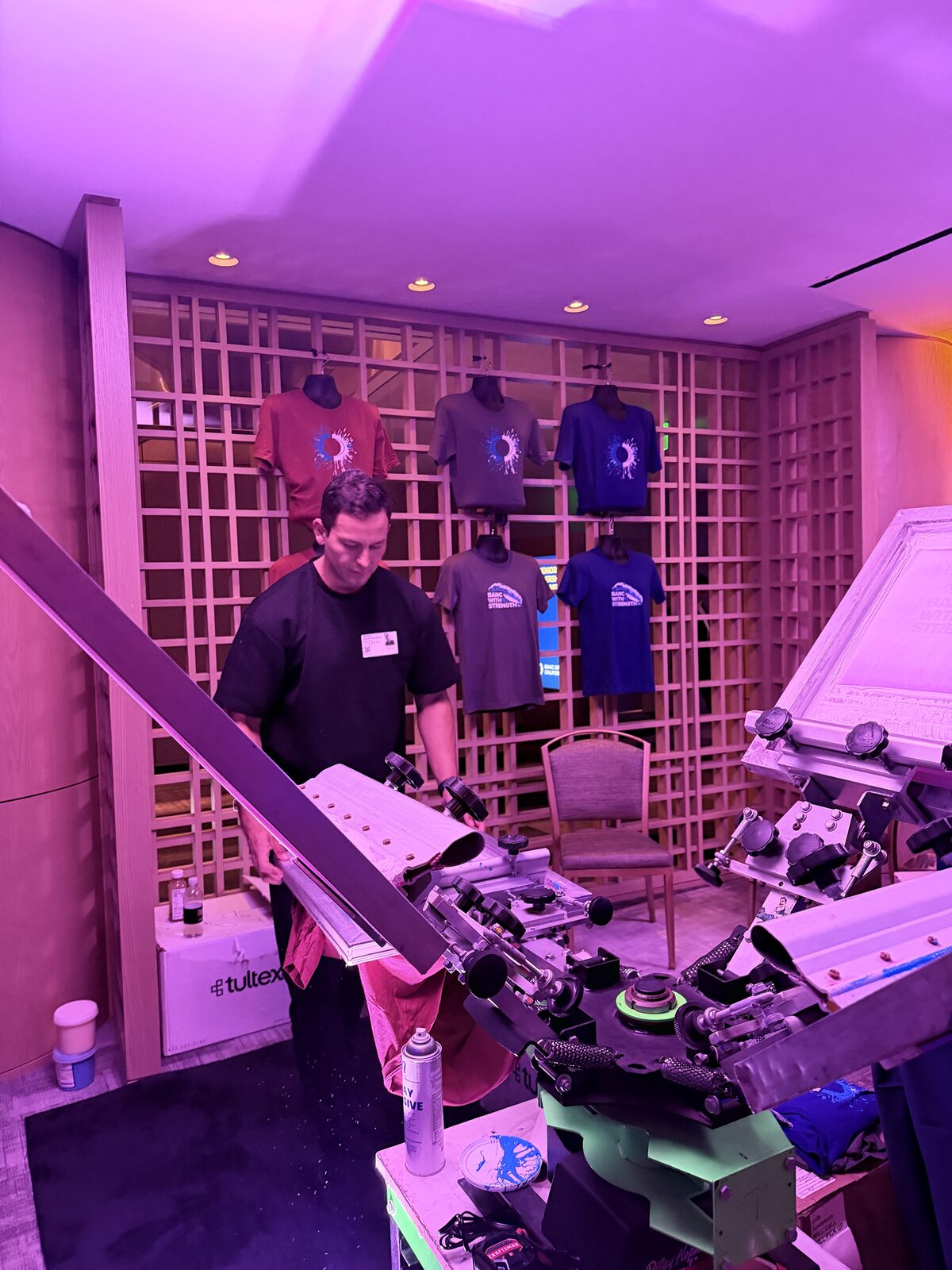 Operator working a live shirt press beside a display wall of finished designs